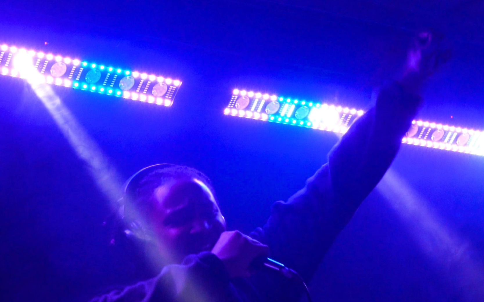 Singer performing on stage under purple-blue lights, holding a microphone with one arm raised.
