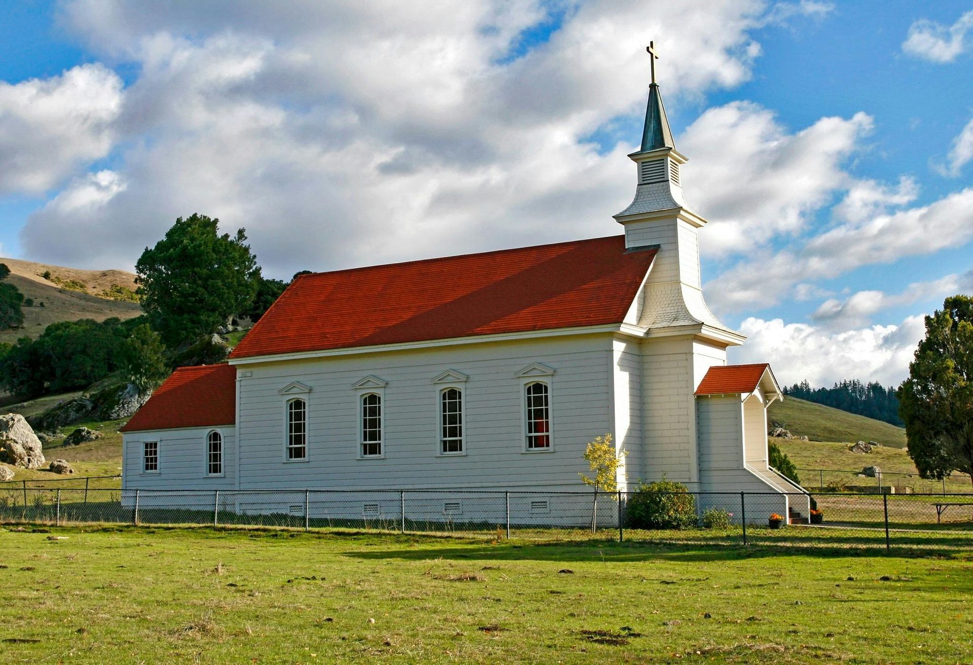 White church with red roof and steeple, set in a green field with hills under a blue, cloudy sky.