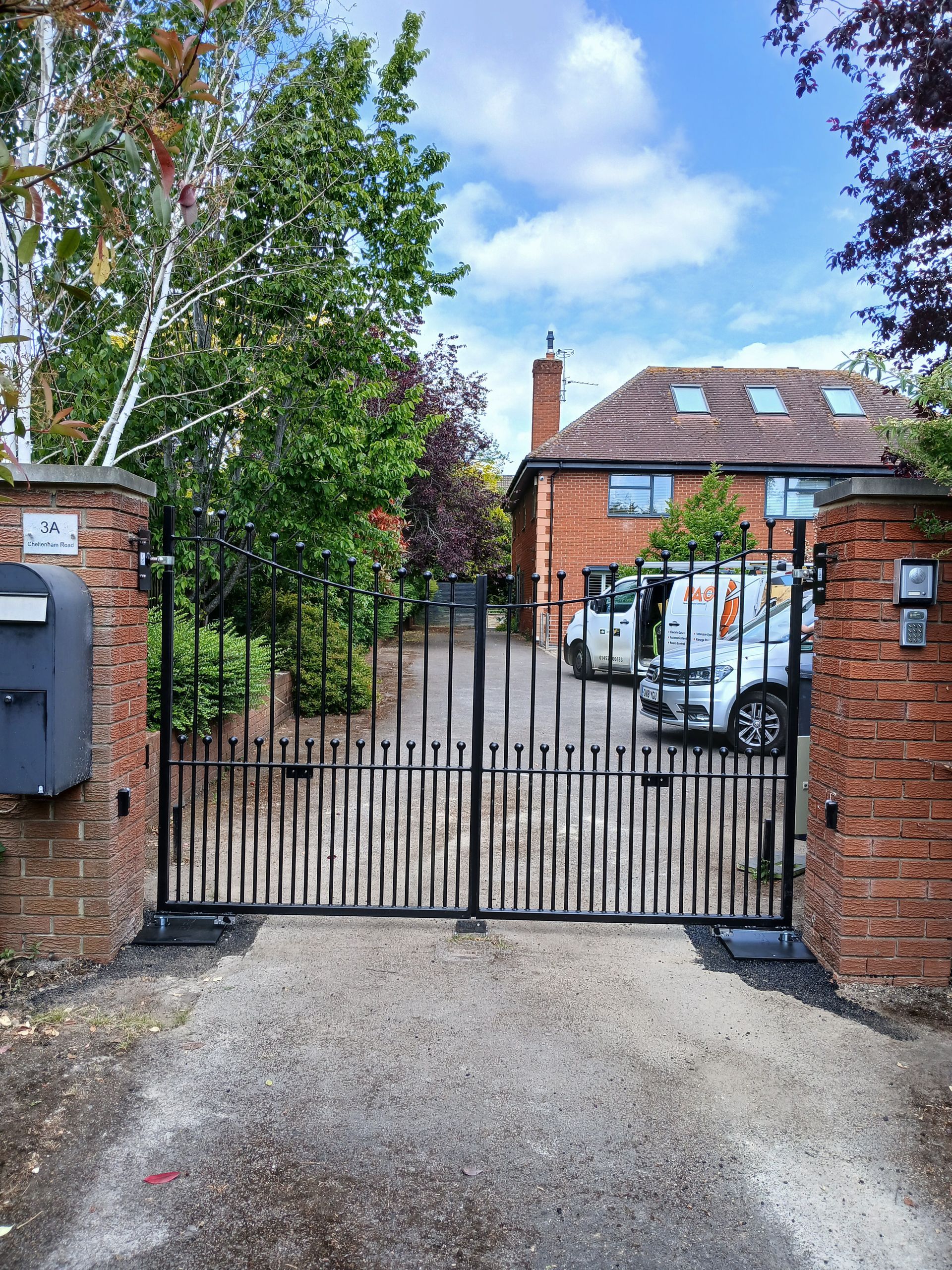 Black iron driveway gates leading to a brick house with a white van parked in the driveway.