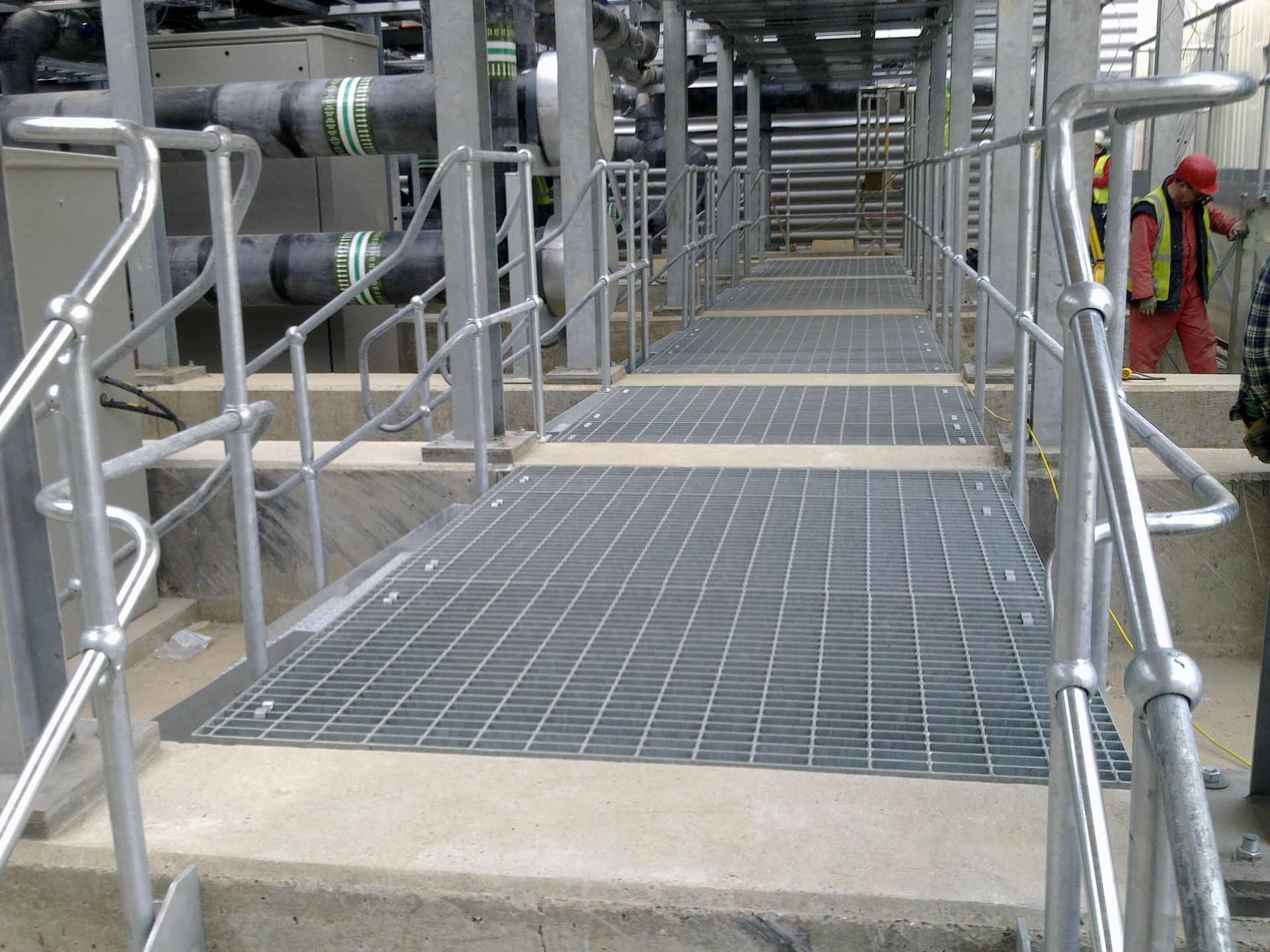 Metal grated walkway with handrails; industrial setting. A worker in the background.