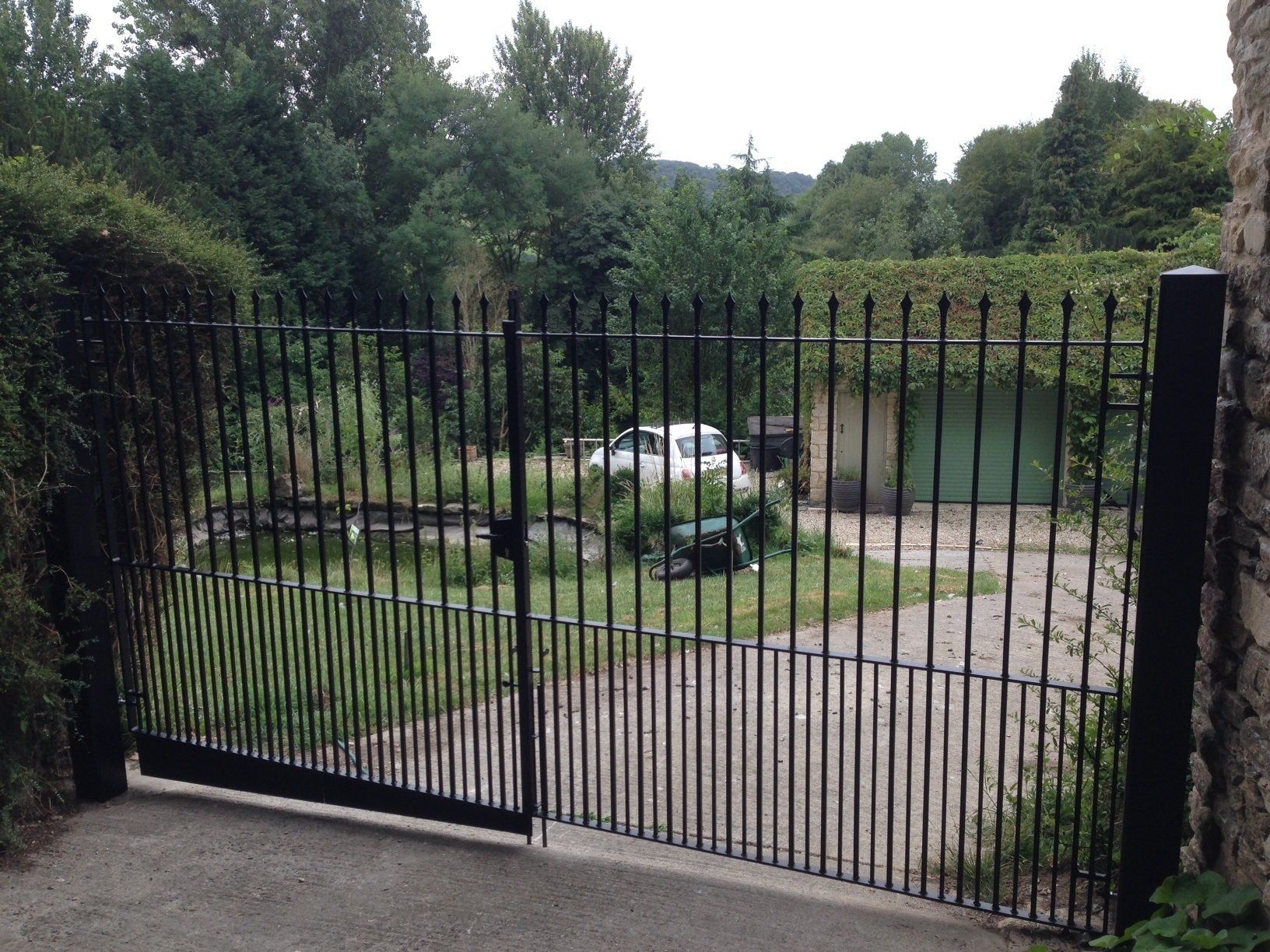 Black metal gate open to a driveway with a car, surrounded by greenery.