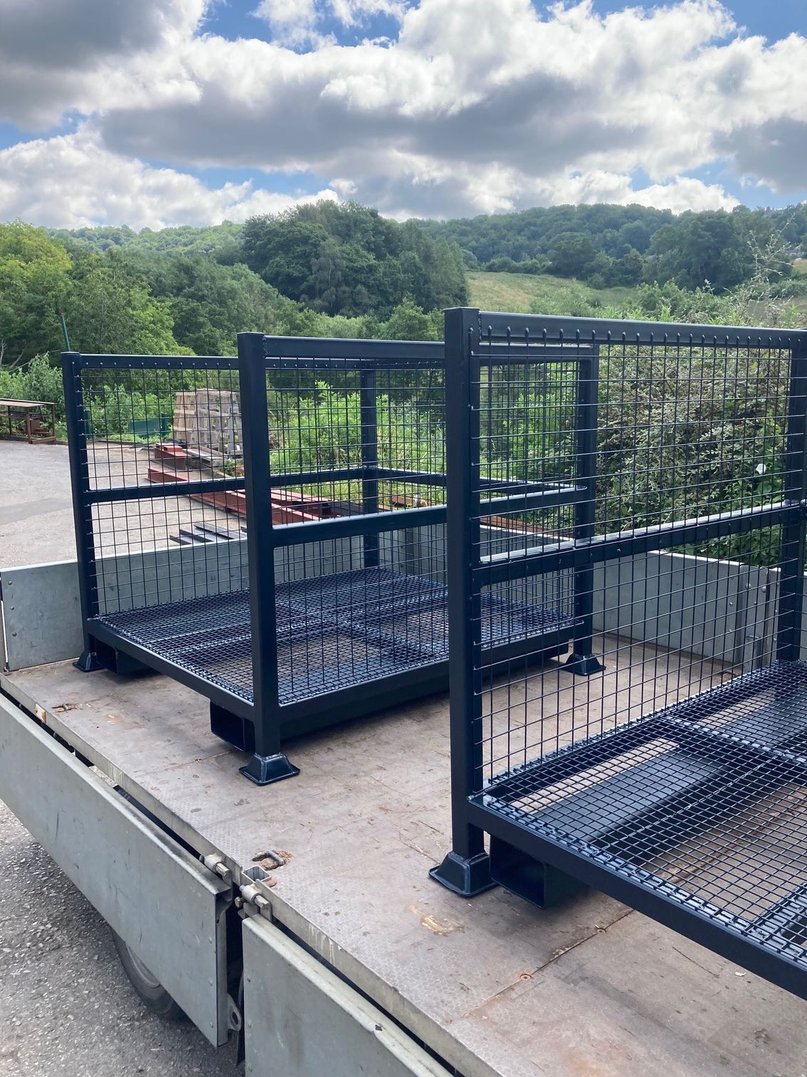 Two dark metal storage racks on the bed of a truck, set against a backdrop of green hills and sky.