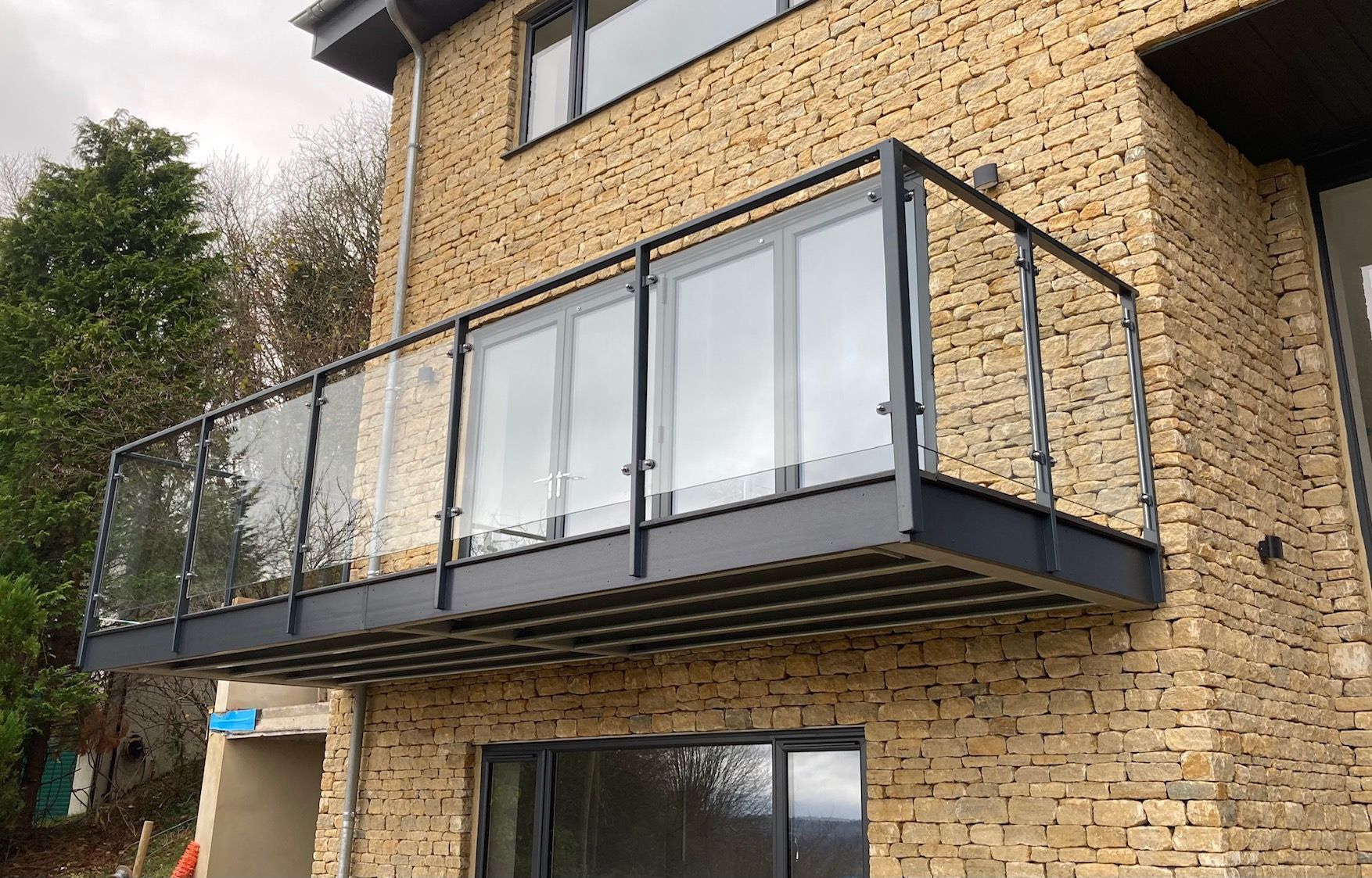 A modern balcony with glass railings and dark gray metal frame, attached to a stone-walled building.