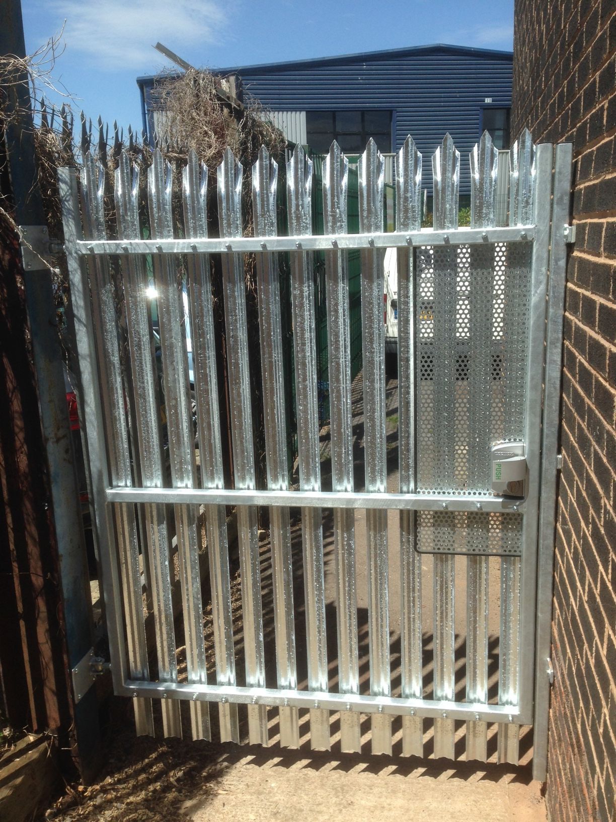 Metal security gate with sharp vertical spikes and perforated panel, set between brick and metal structures.
