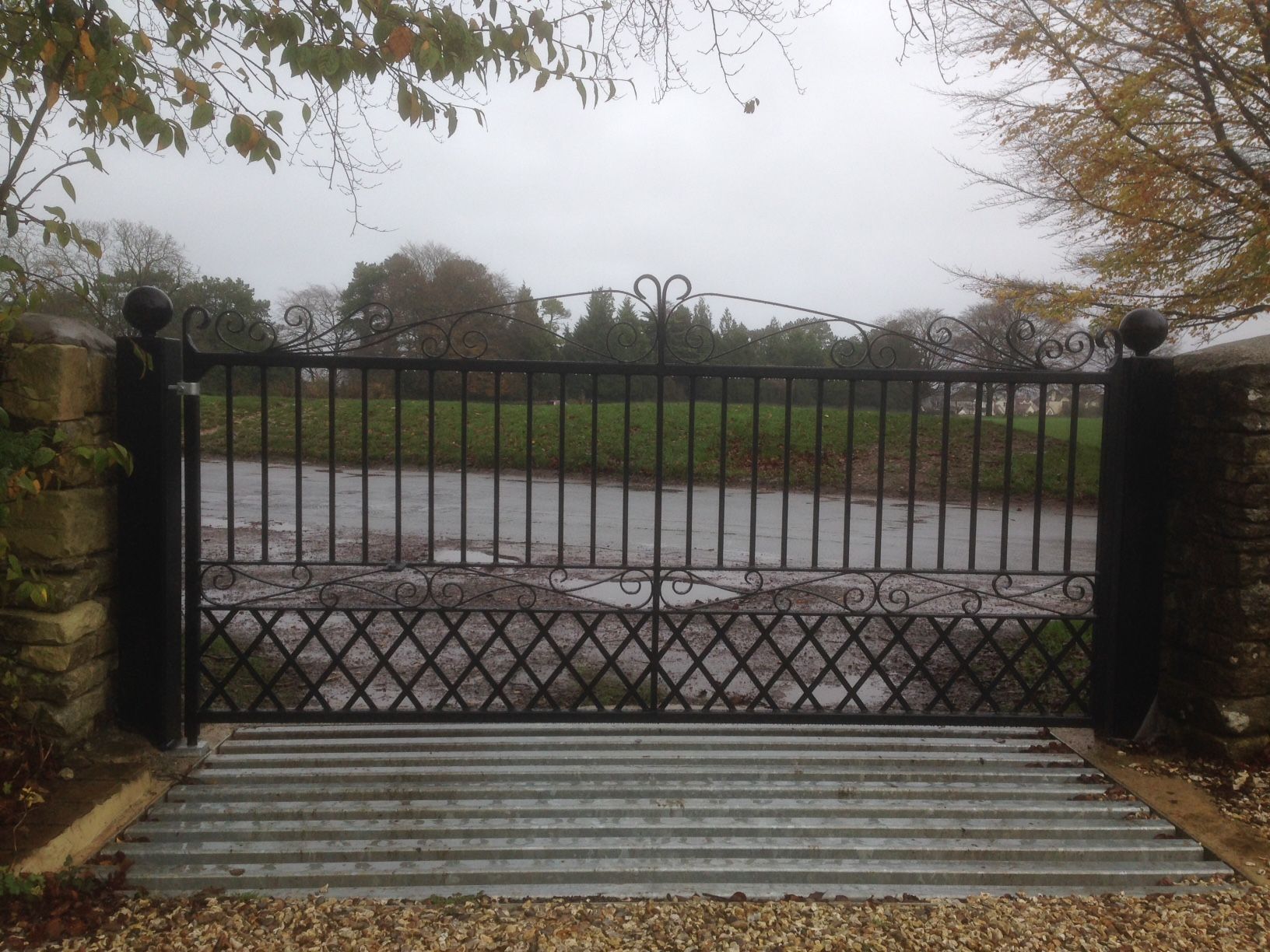 Black metal gate with decorative top, over a cattle grid, in a rural setting.