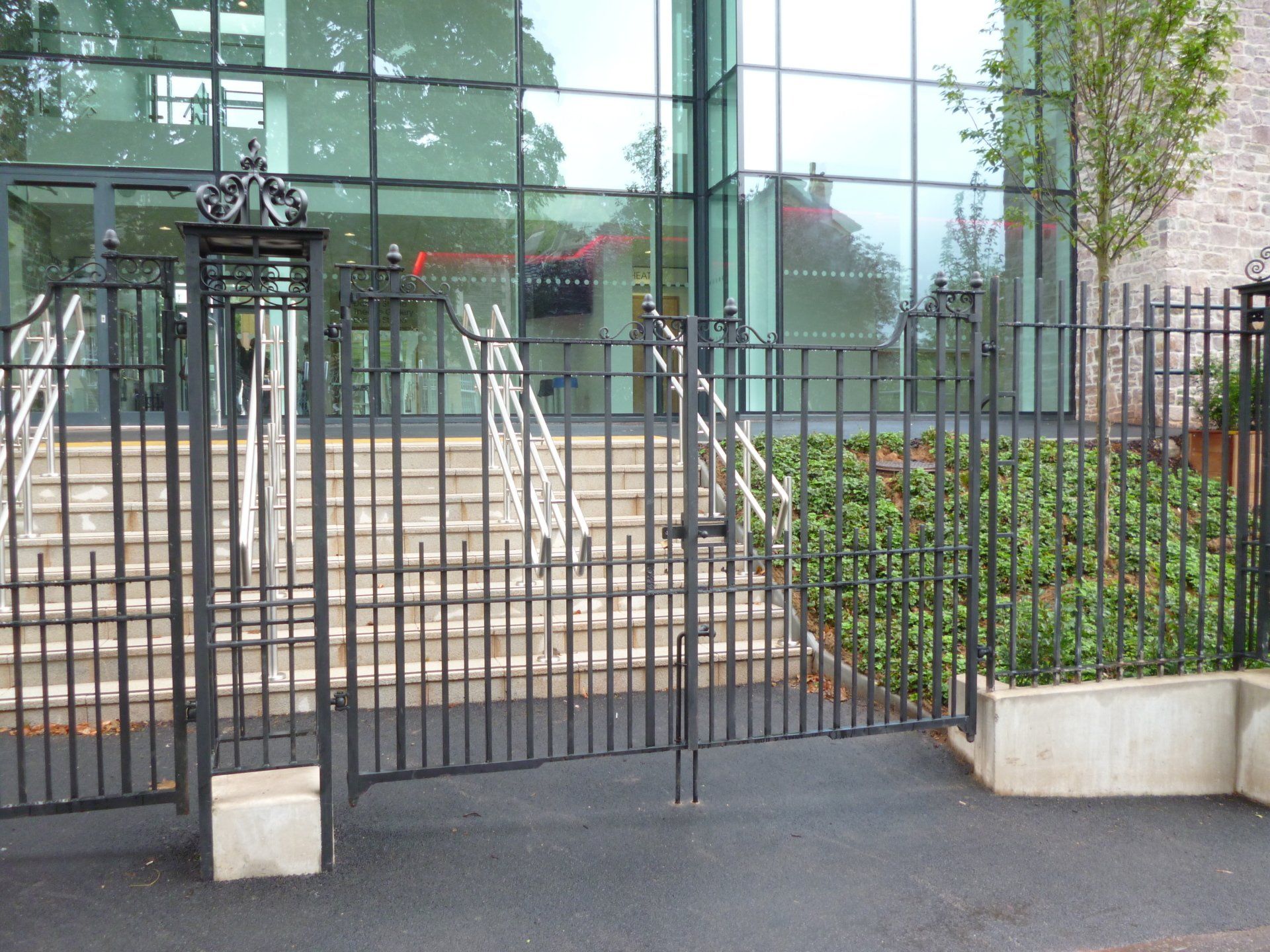Black metal gate and fence in front of a modern building with glass windows, steps and greenery.