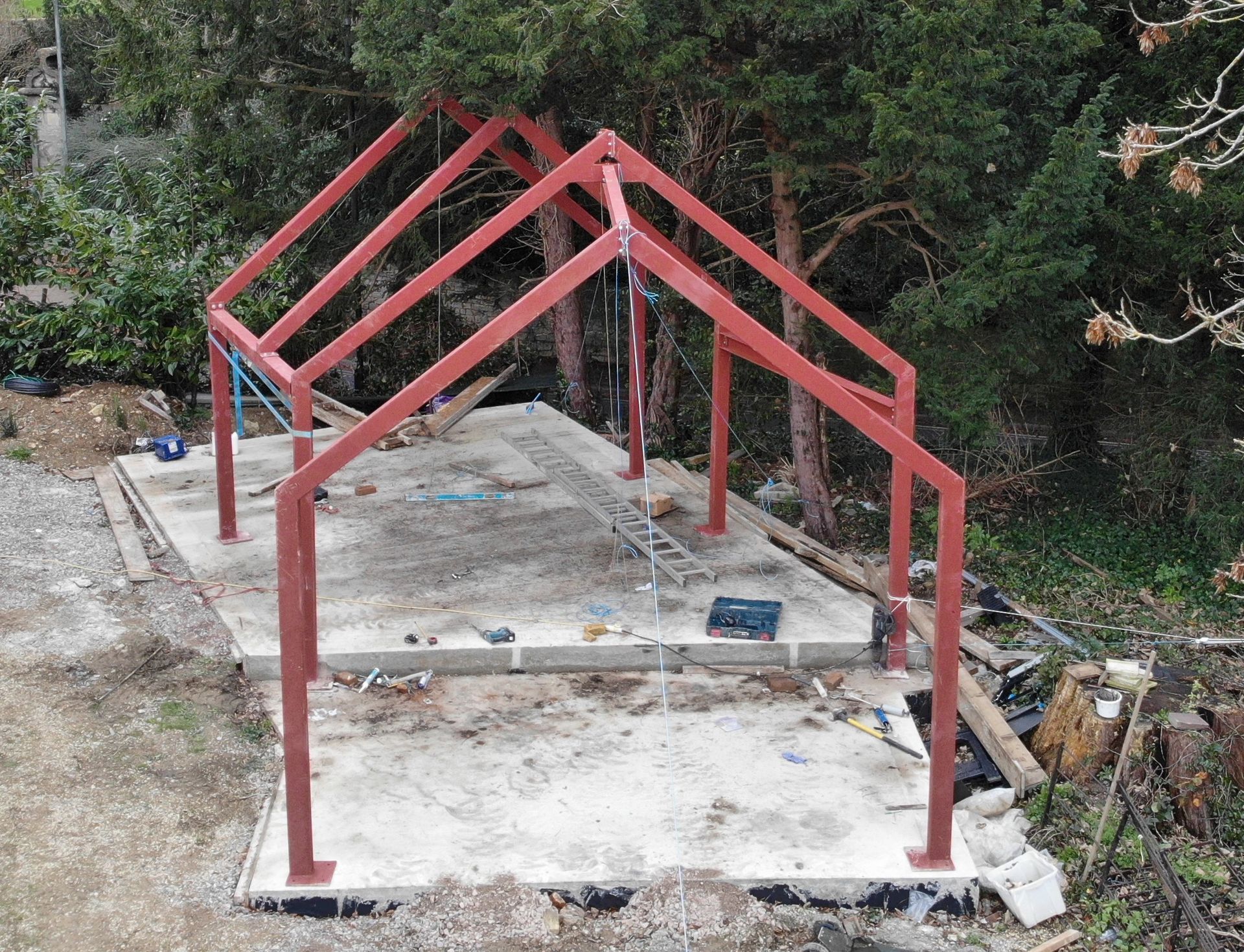 Red metal frame structure on a concrete slab, with surrounding trees. Construction site.