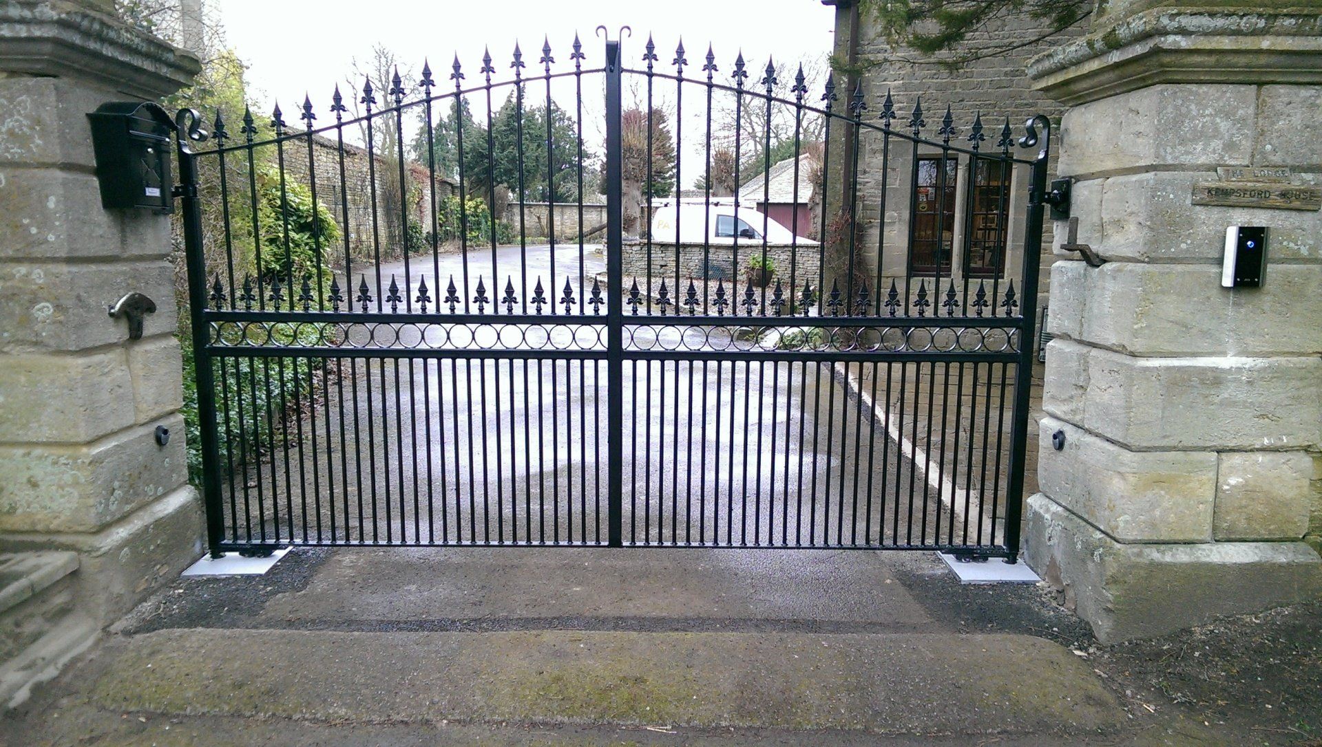 Black wrought-iron gate between stone pillars, leading to a driveway, with a security system.