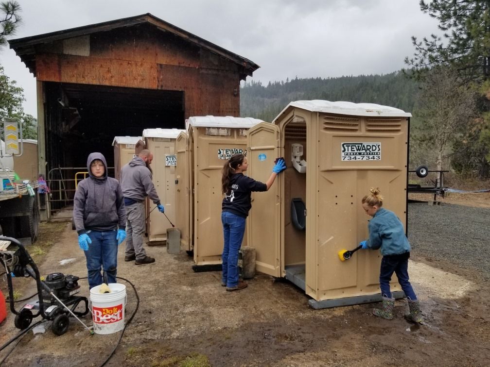 People cleaning portable toilets in front of a wooden structure under a cloudy sky.