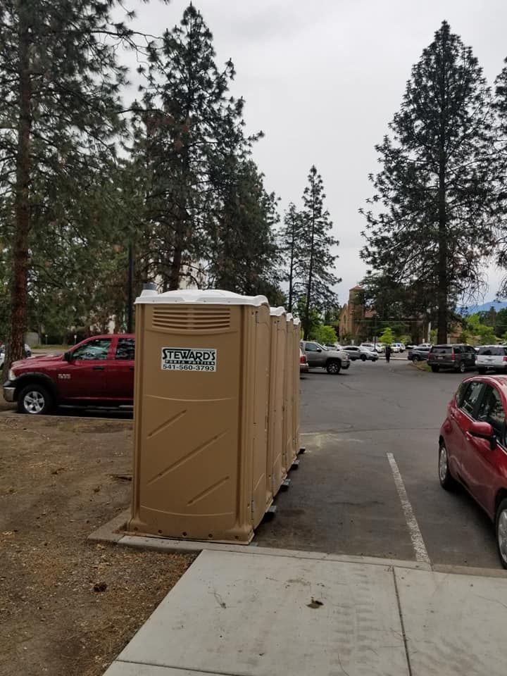 Row of tan portable toilets in a parking lot, trees in the background, overcast sky.