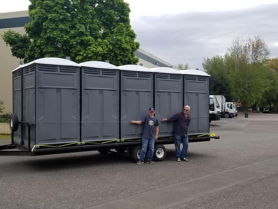 Two men stand beside a trailer carrying five gray portable toilets, parked on asphalt.