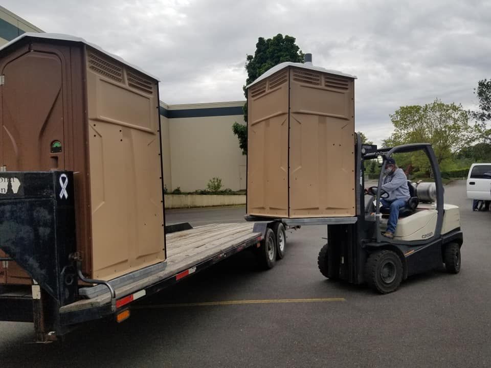 Forklift operator moving a portable toilet onto a trailer with another toilet. Outdoors, day.