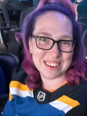 Smiling person with purple hair and glasses wearing a blue, yellow, and white hockey jersey