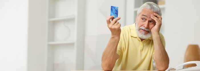 An elderly man is sitting at a table holding a credit card wondering What is Credit Card Fraud.