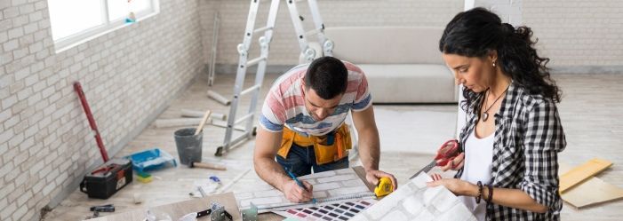Image of two people in a home under renovation, smiling amidst a messy background with tools and materials, indicating ongoing upgrades like energy-efficient appliances and modern lighting, reflecting the process of enhancing their living space.
