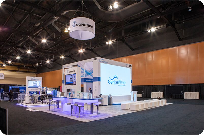 Trade show booth with white and blue displays and a large screen.