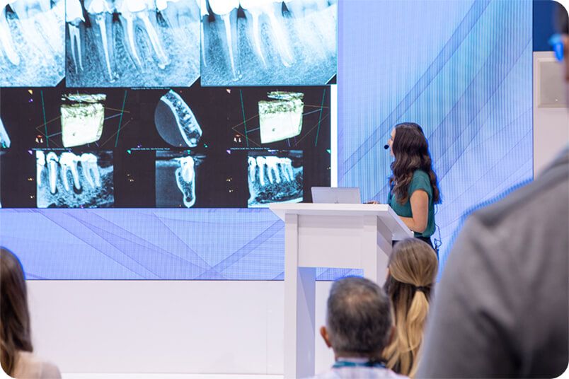 Woman giving presentation on dental x-rays. Audience watches; large screen displays various x-ray images.