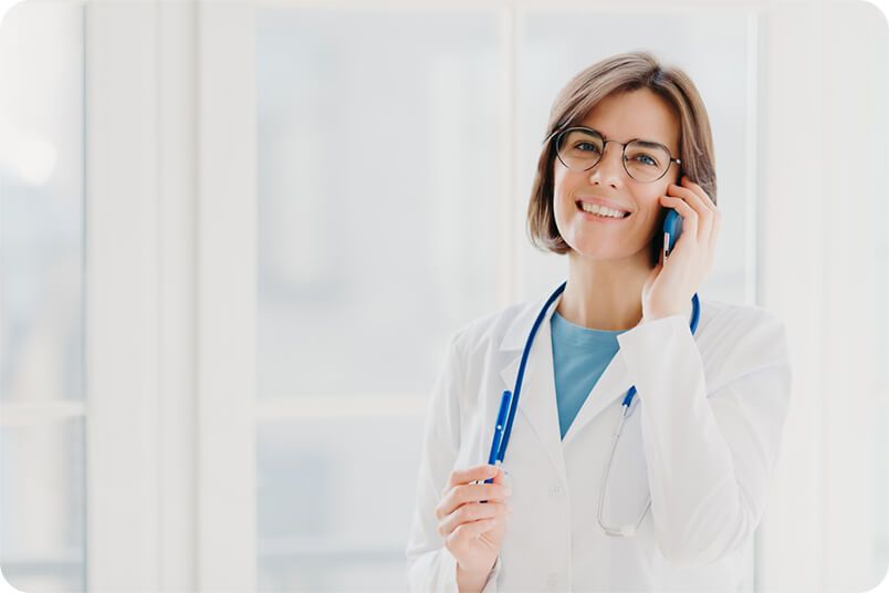 Doctor wearing a white coat and glasses smiles while talking on a phone, stethoscope around her neck.