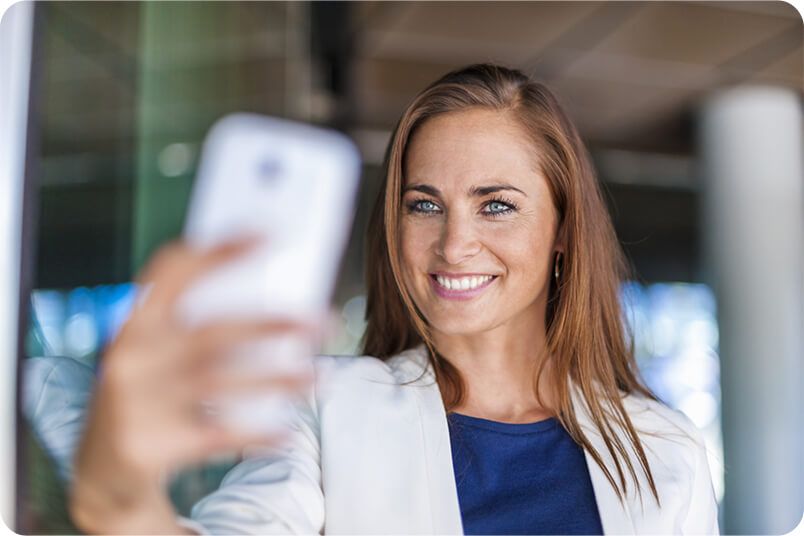 Woman taking a selfie with a smartphone, smiling. Wearing a white jacket and blue top, in a building.