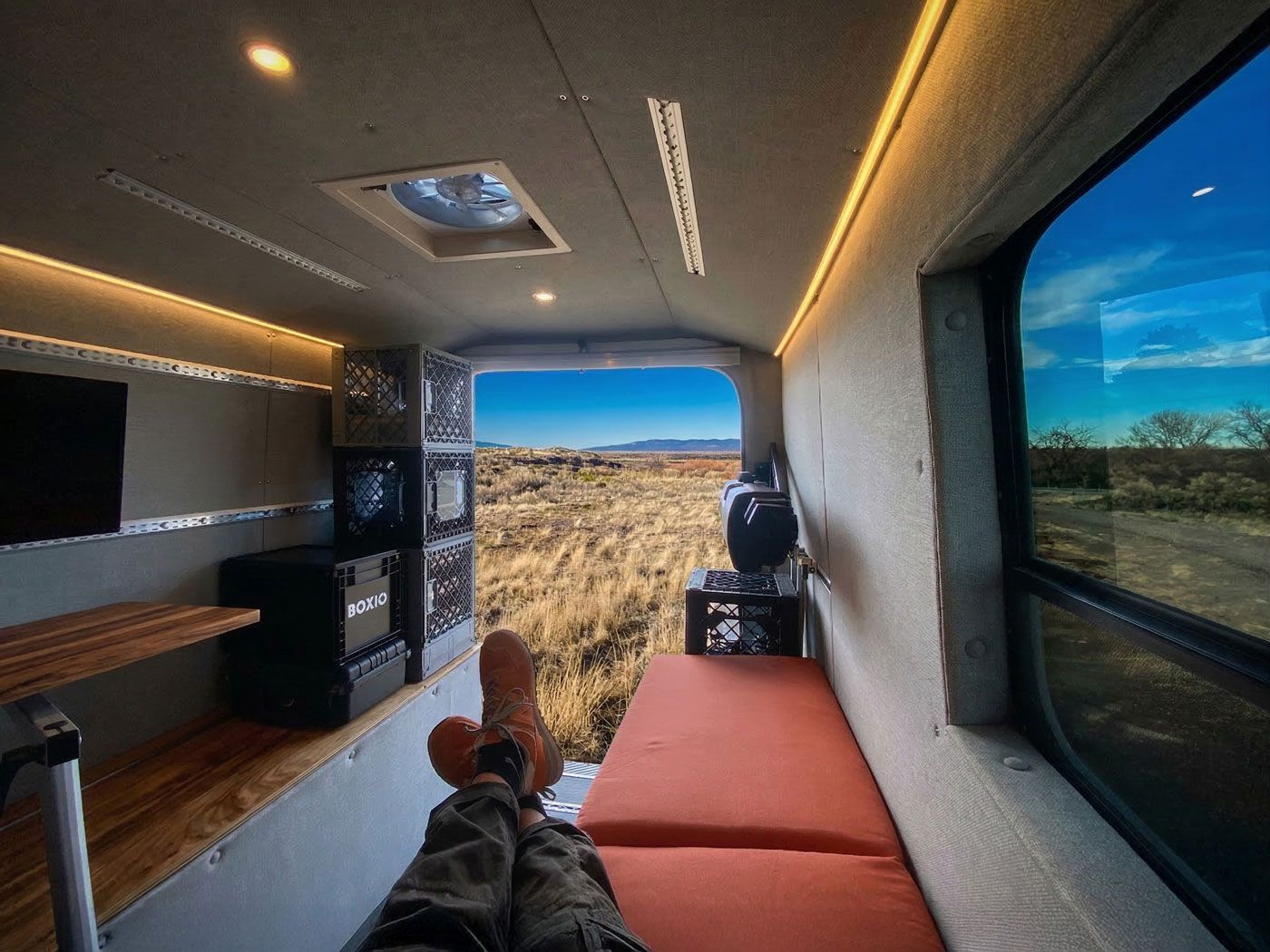 Inside a camper van, a person lies on a bed facing the open rear doors and desert landscape.