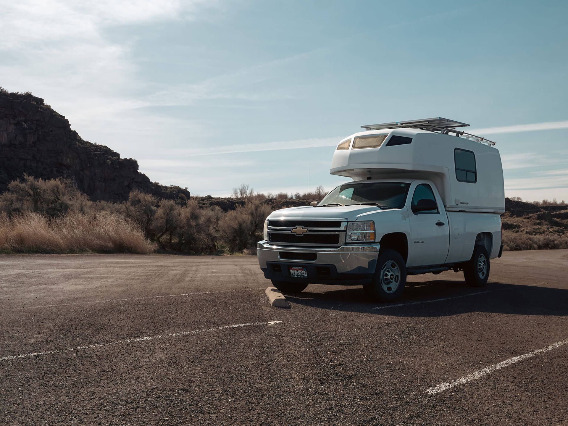 White pickup truck with camper shell parked on an empty paved lot near rocky hills under a clear sky
