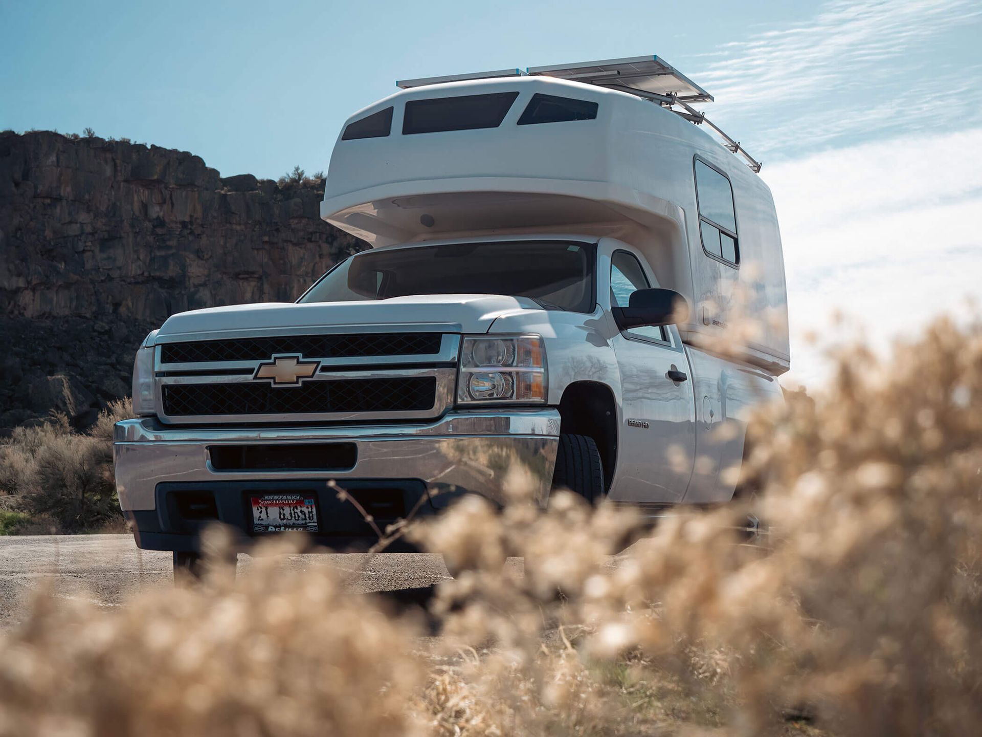 White pickup truck with camper parked on a dusty road near rocky cliffs under a clear sky