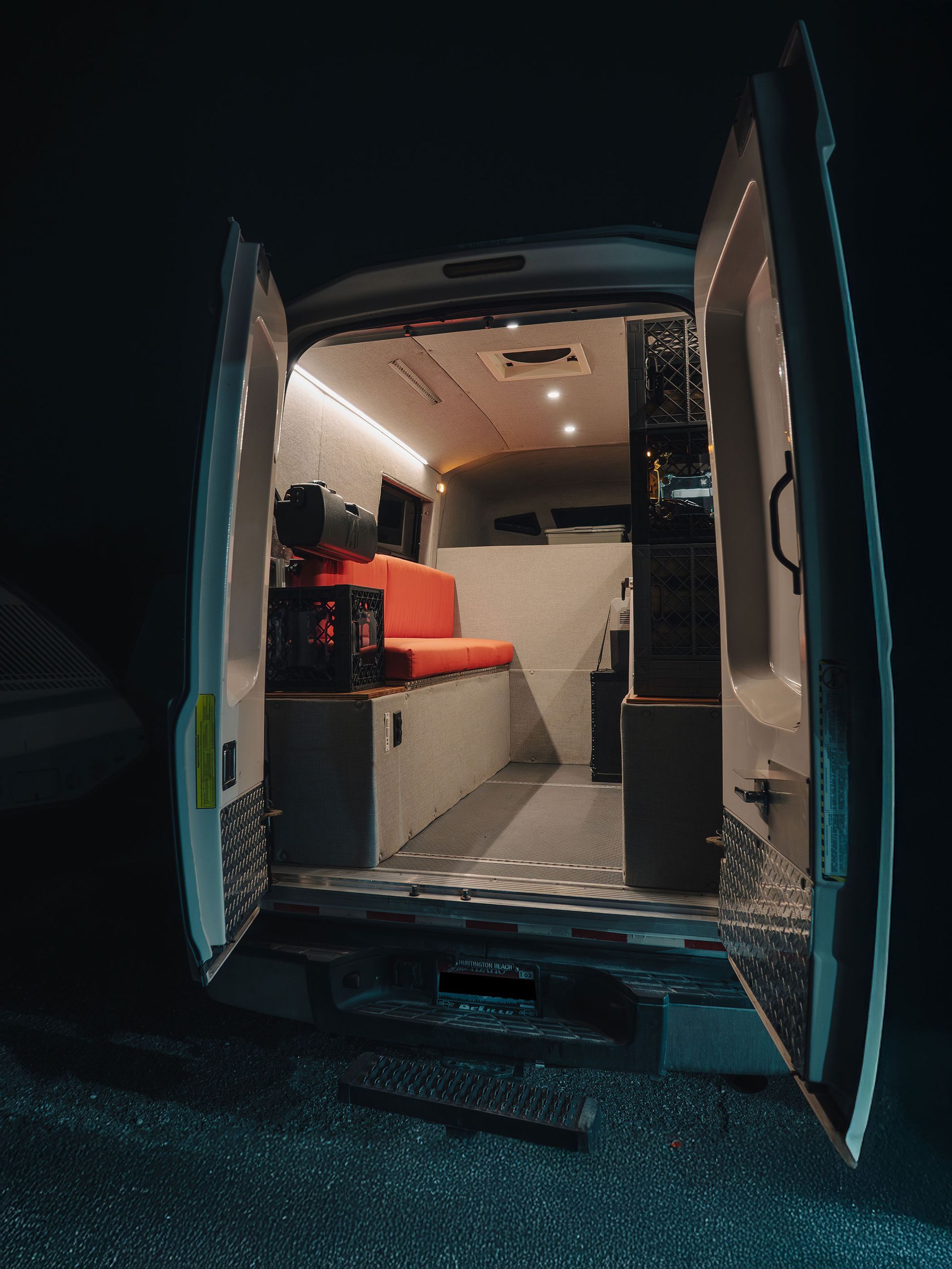 Open van door to a compact camper van interior with red bench seating and white cabinets at night