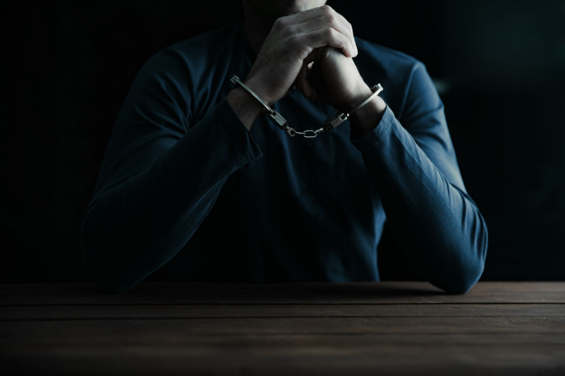 Person in handcuffs, hands clasped, sitting at a wooden table in a dimly lit setting.