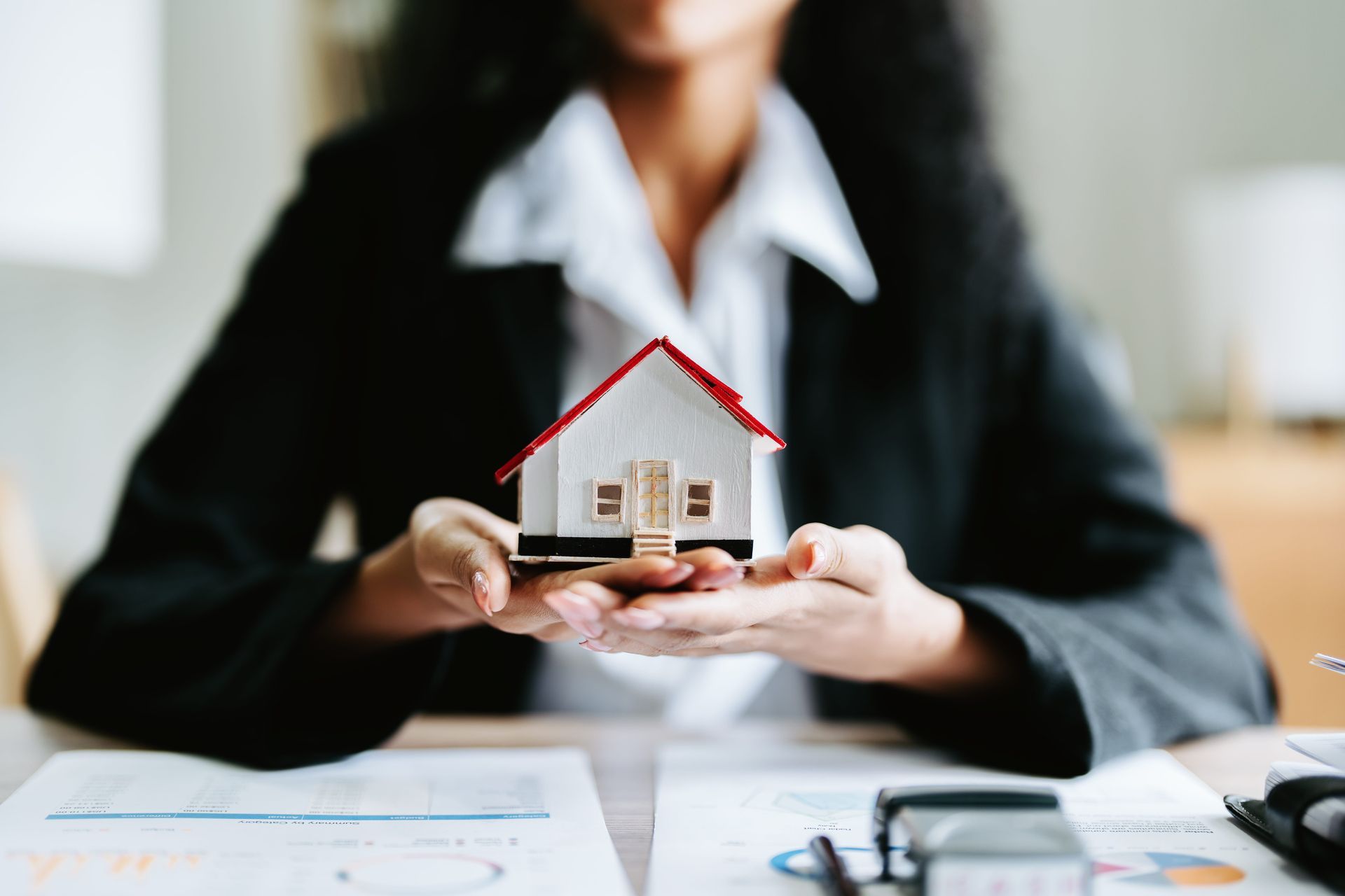 Person holding a small house model at a desk with paperwork, suggesting real estate or home insurance.