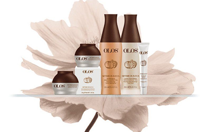 Un set di prodotti per la cura della pelle OLAS color beige, composto da vasetti, flaconi e tubetti su una superficie bianca con un delicato sfondo a petali.