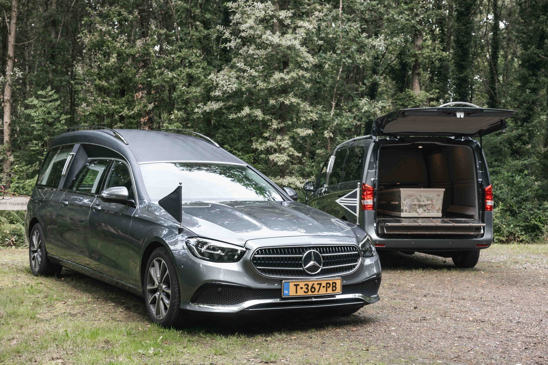 Een zwarte Mercedes-bus en stationwagen staan geparkeerd op een onverharde bosweg,