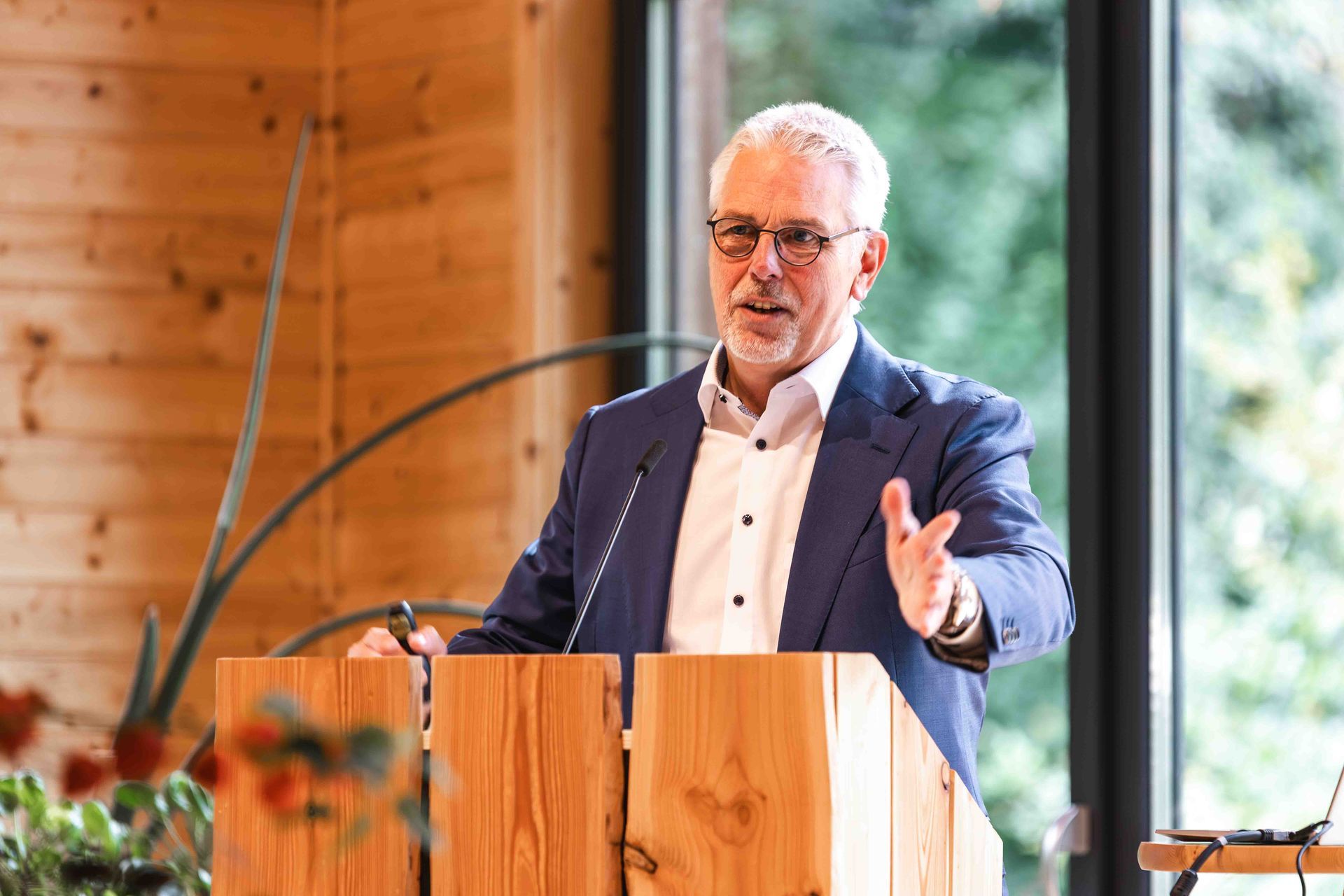 Een man spreekt vanaf een houten podium in een met houten panelen beklede ruimte en gebaart tijdens een presentatie.