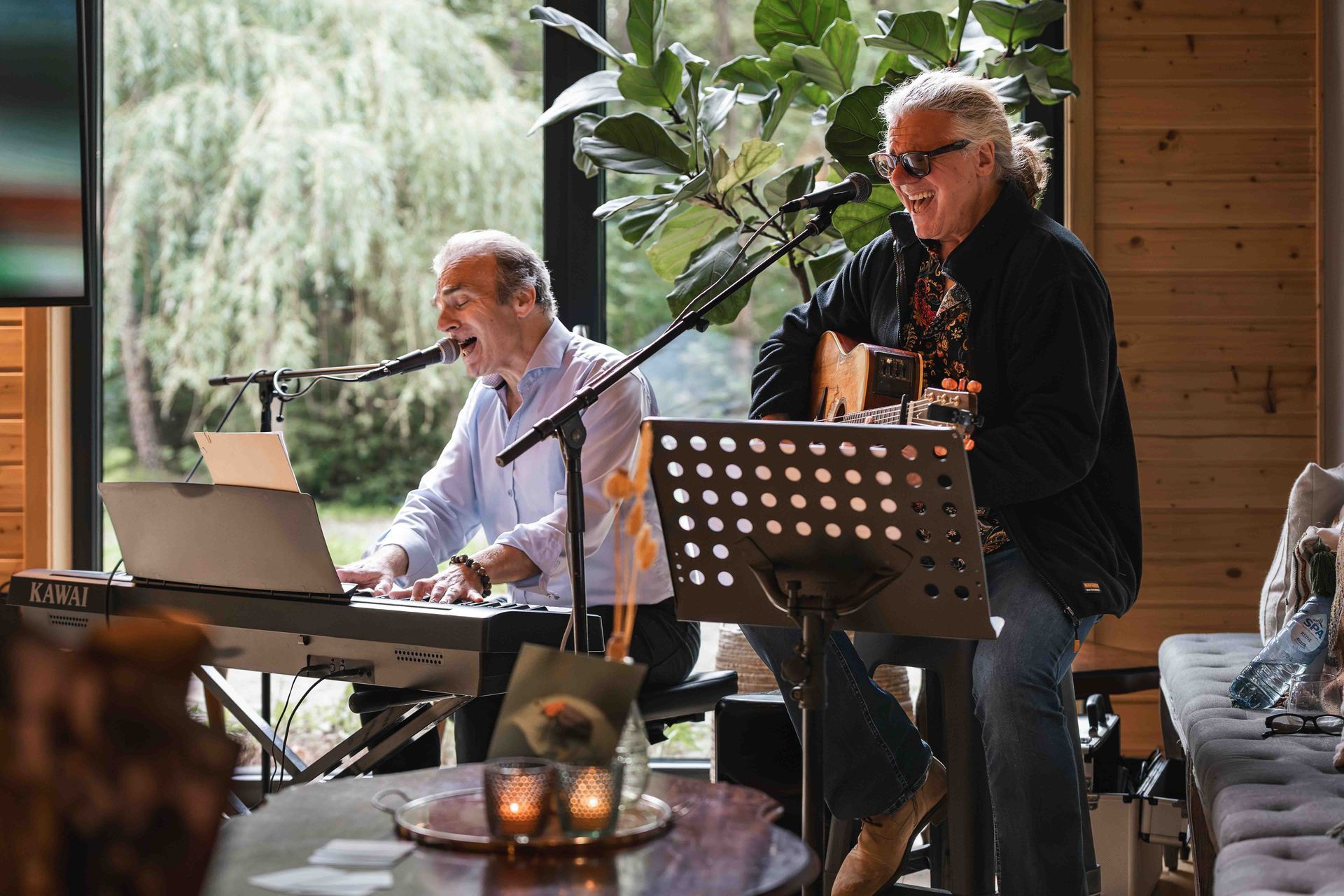 Twee muzikanten treden binnen op bij een raam, de een achter een keyboard en de ander zingt in een microfoon.