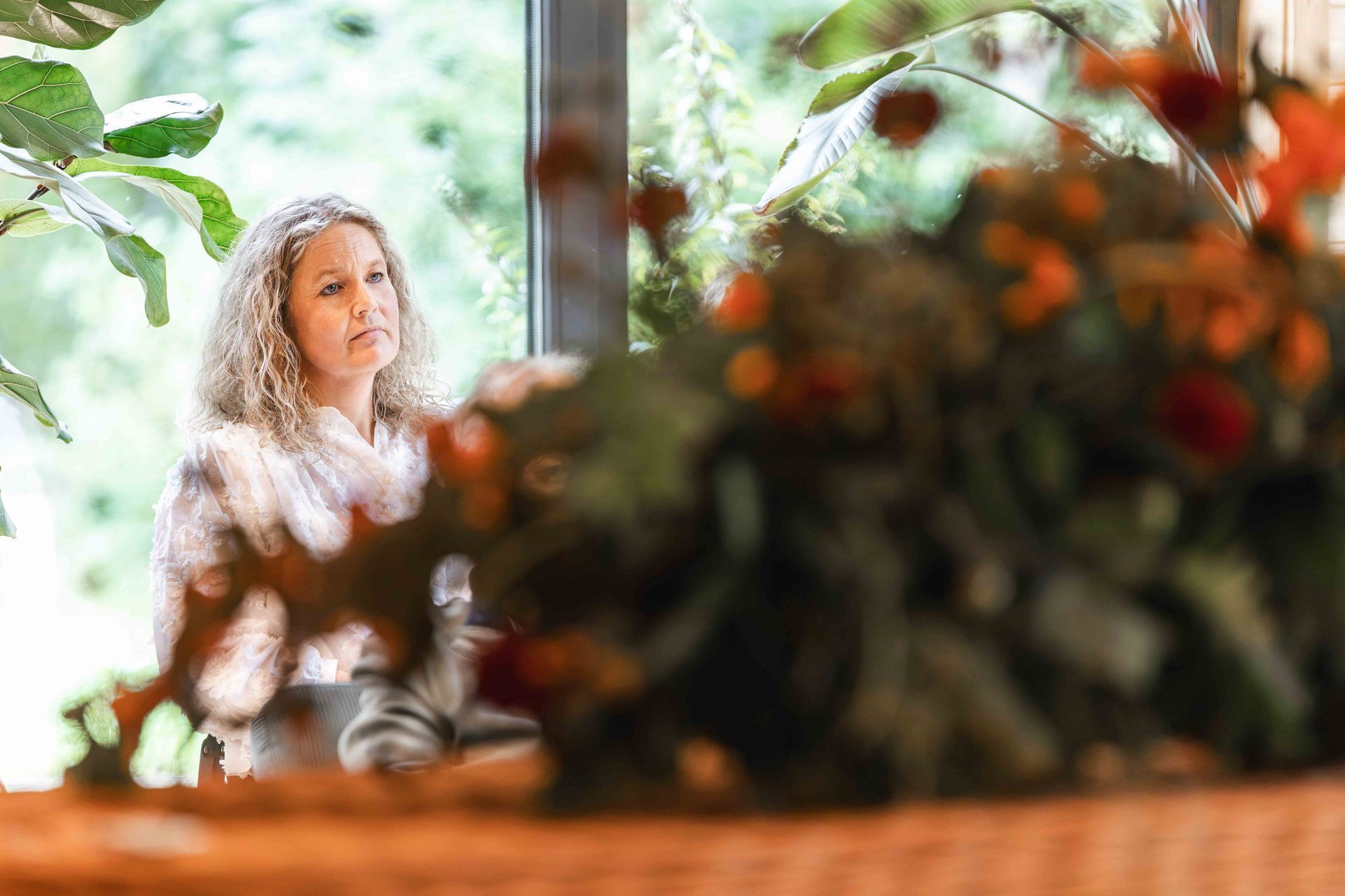 Vrouw zit bij een licht raam, gedeeltelijk bedekt door bloemen en bladeren op de voorgrond.