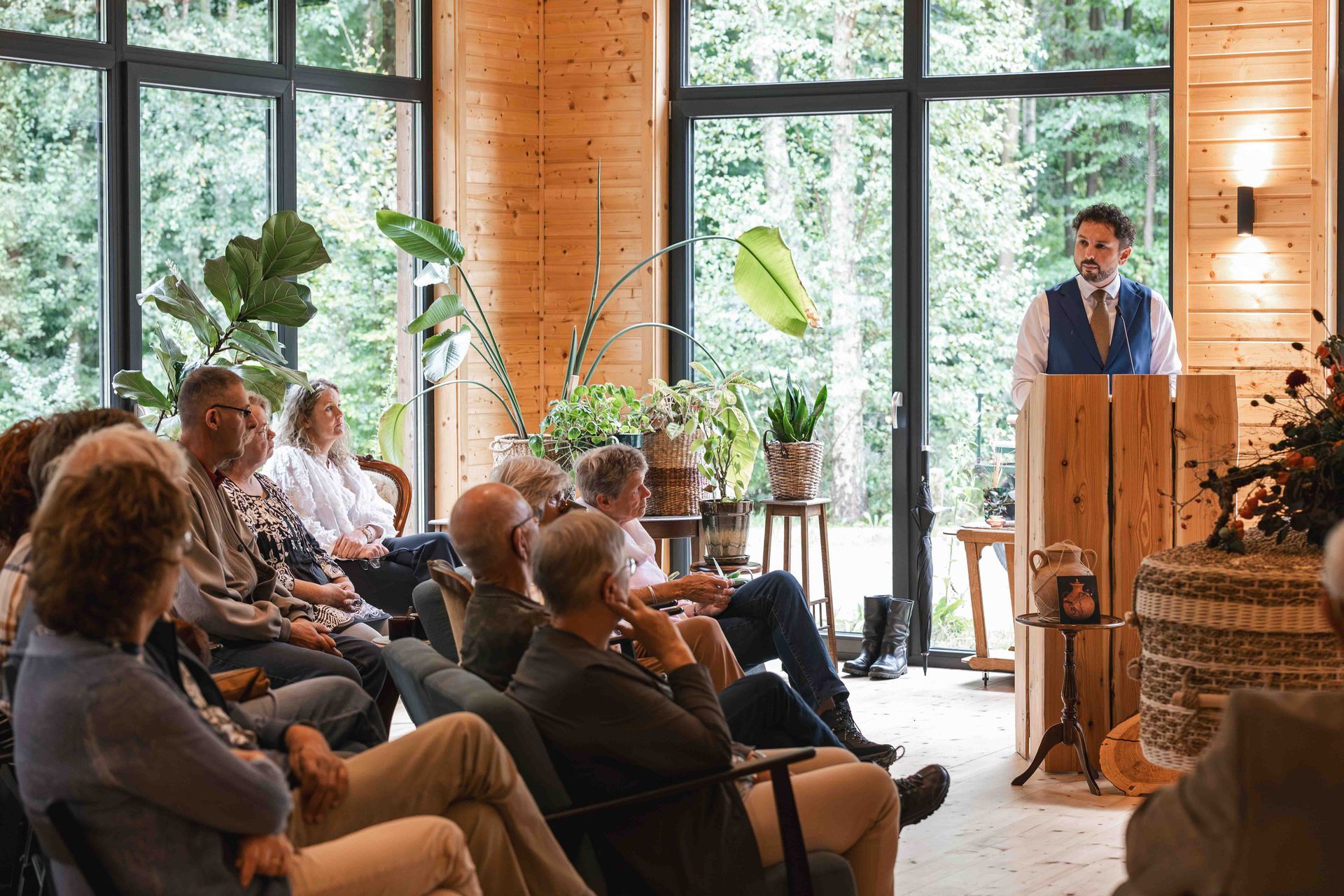 Het publiek luistert naar een spreker vanaf een houten podium in een lichte, met planten gevulde ruimte.