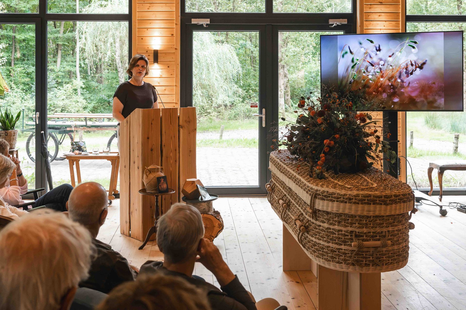 Spreker op een podium in een lichte zaal, met een groot scherm en een rieten bloemstuk met bloemen als achtergrond.
