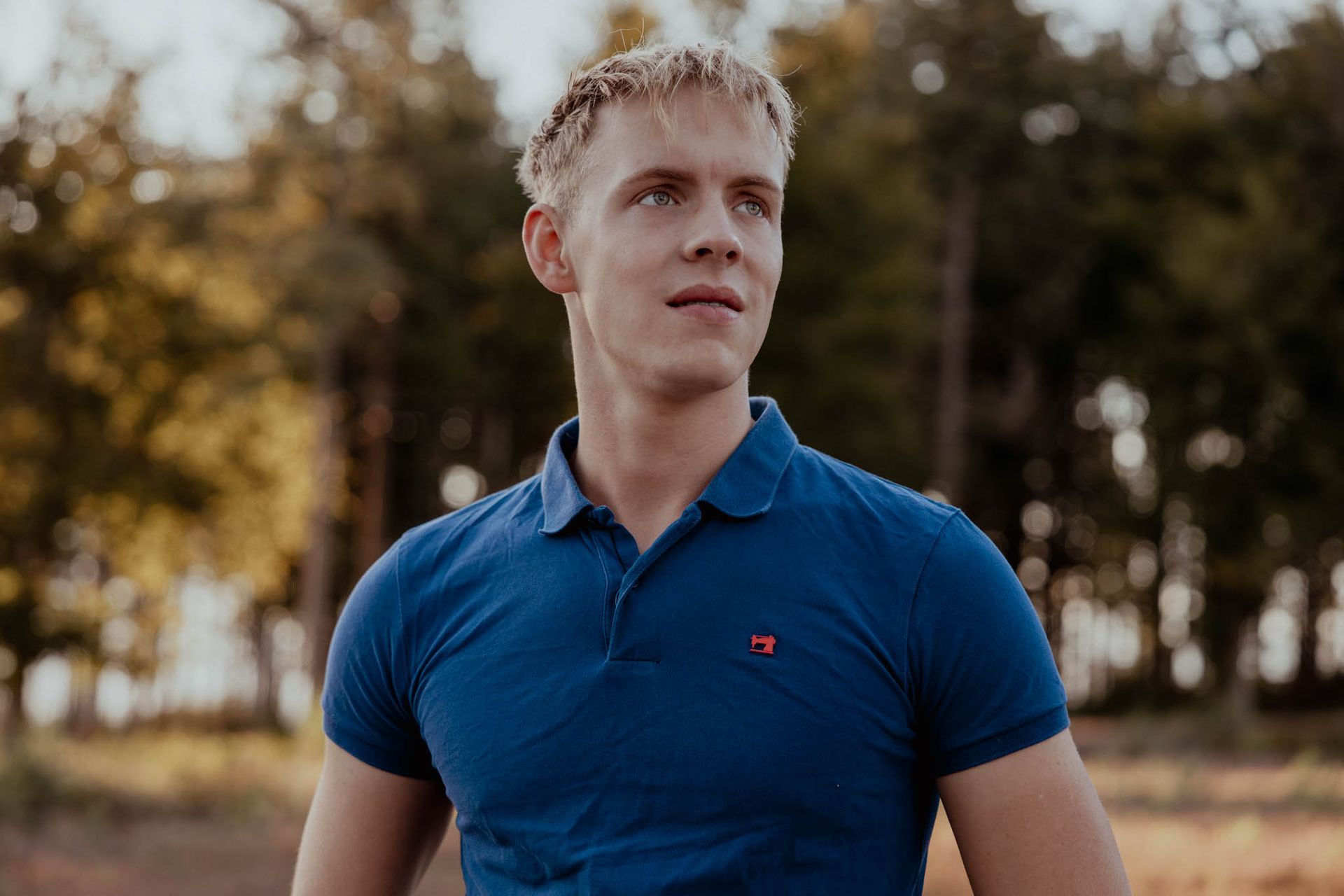 Een blondharige persoon in een blauw poloshirt kijkt weg van de camera in een park, met bomen op de achtergrond.