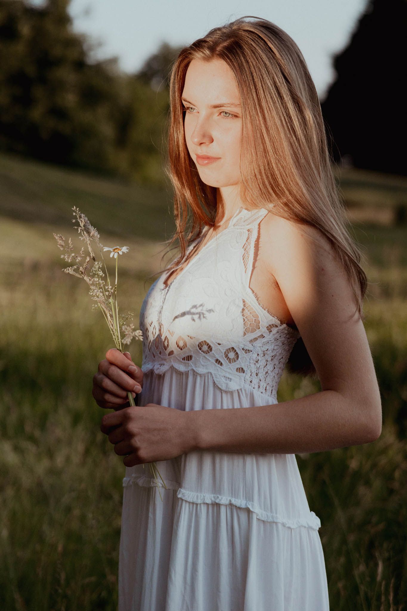 Een vrouw in een witte jurk houdt bloemen vast in een zonovergoten veld en kijkt niet naar de camera.