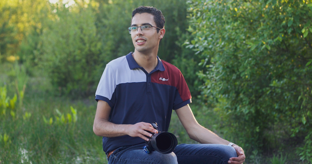 Een man met een camera in zijn hand, gekleed in een poloshirt, zit buiten bij water en groen en glimlacht.