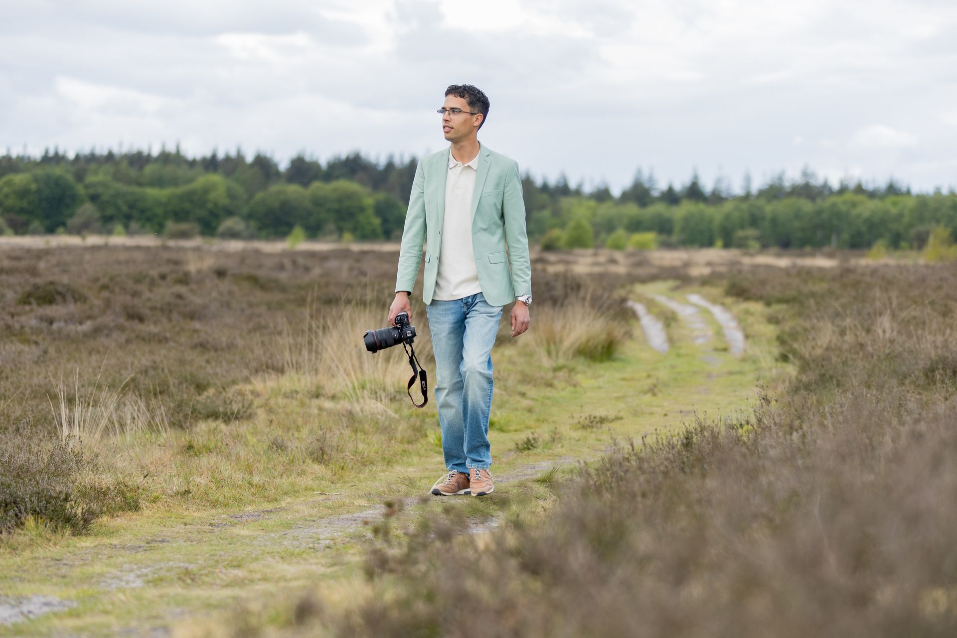 Een man in poloshirt en spijkerbroek zit op een houten bankje, houdt een cameralens vast en kijkt opzij, naar buiten.