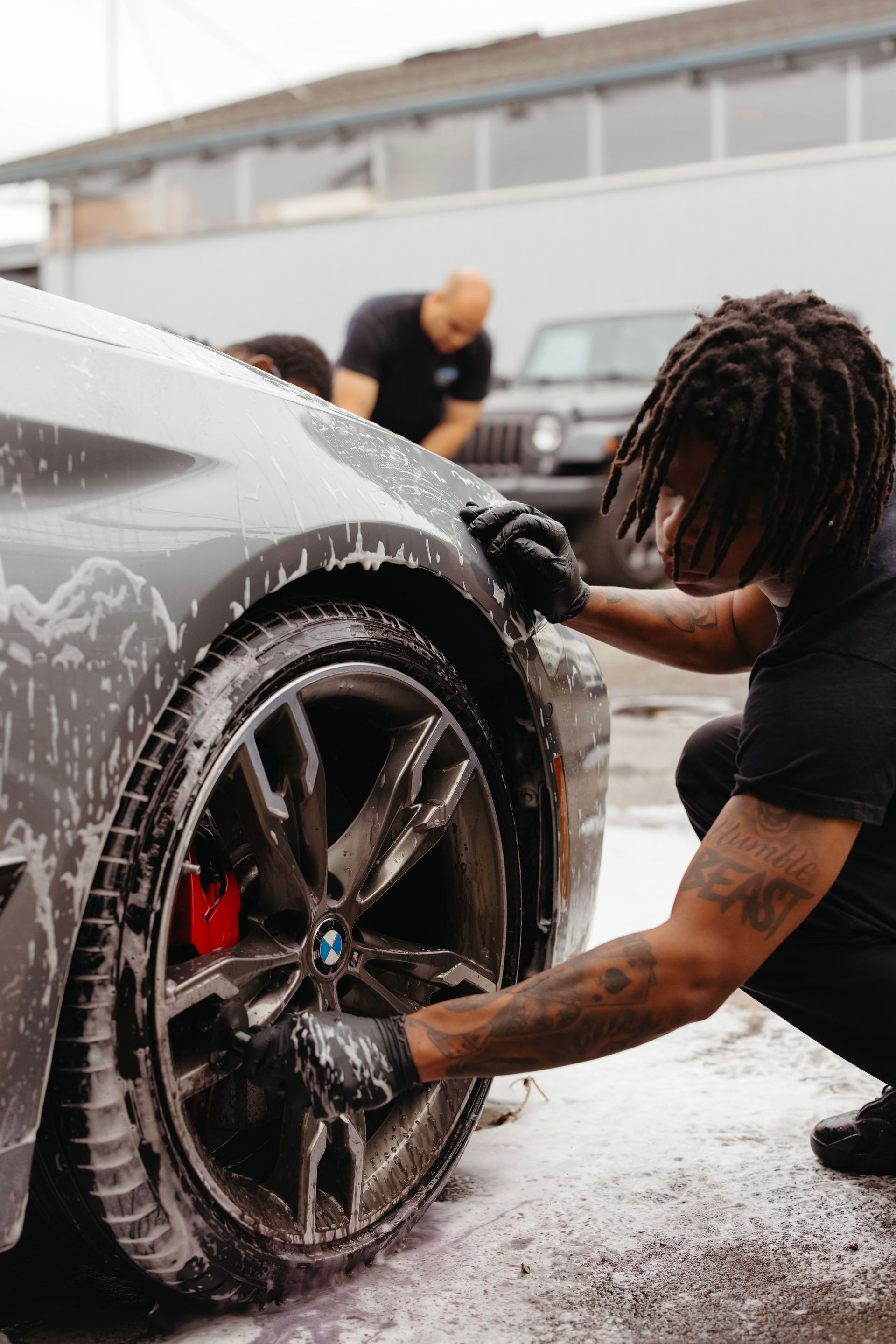 Person washing a black sports car wheel with soap in a wet outdoor car wash area