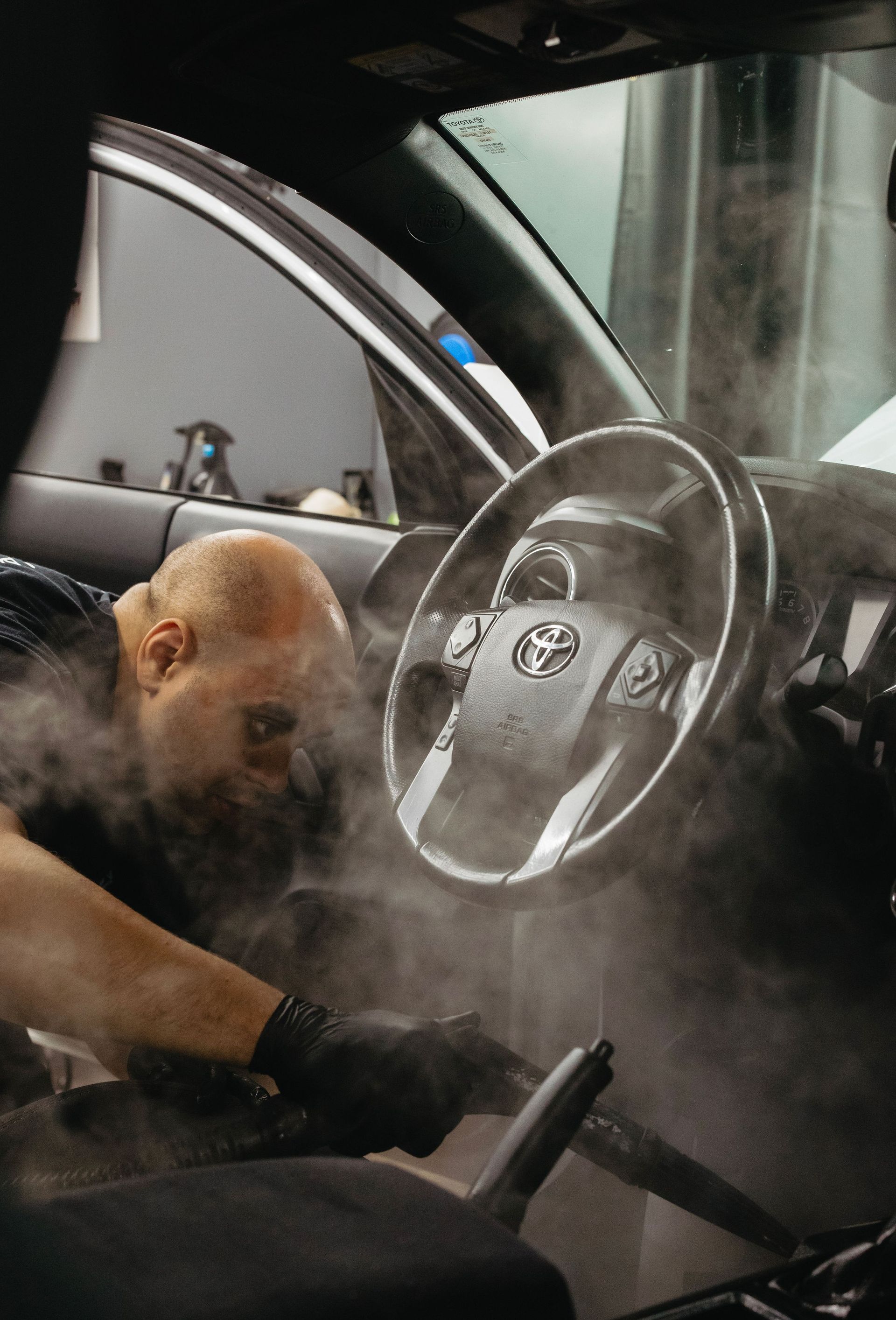 Mechanic cleaning a car interior with steam, door open and steering wheel visible