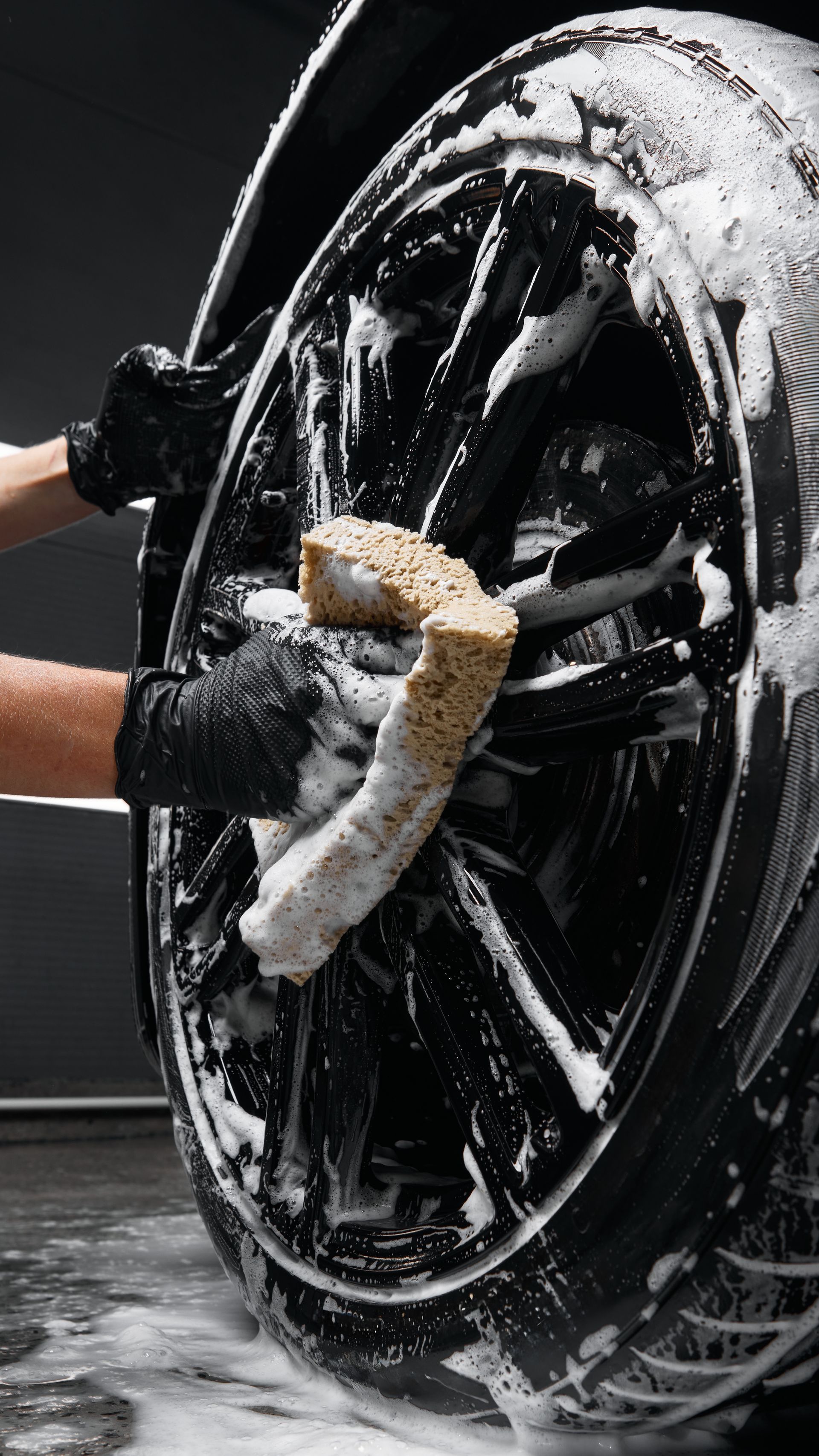 Gloved hands scrub a black car wheel with soap and water in a car wash.
