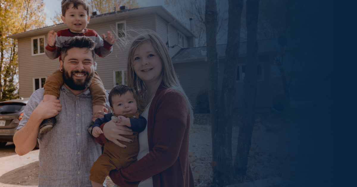 A smiling family poses outdoors with a child sitting on a parent's shoulders and another being held in front.