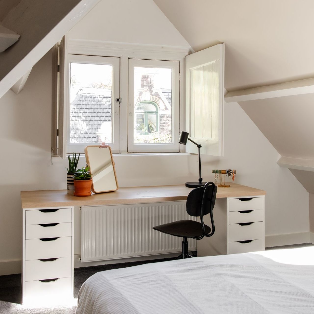 Bureau met raam in een zolderkamer. Witte lades ondersteunen een houten blad met planten en een spiegel.