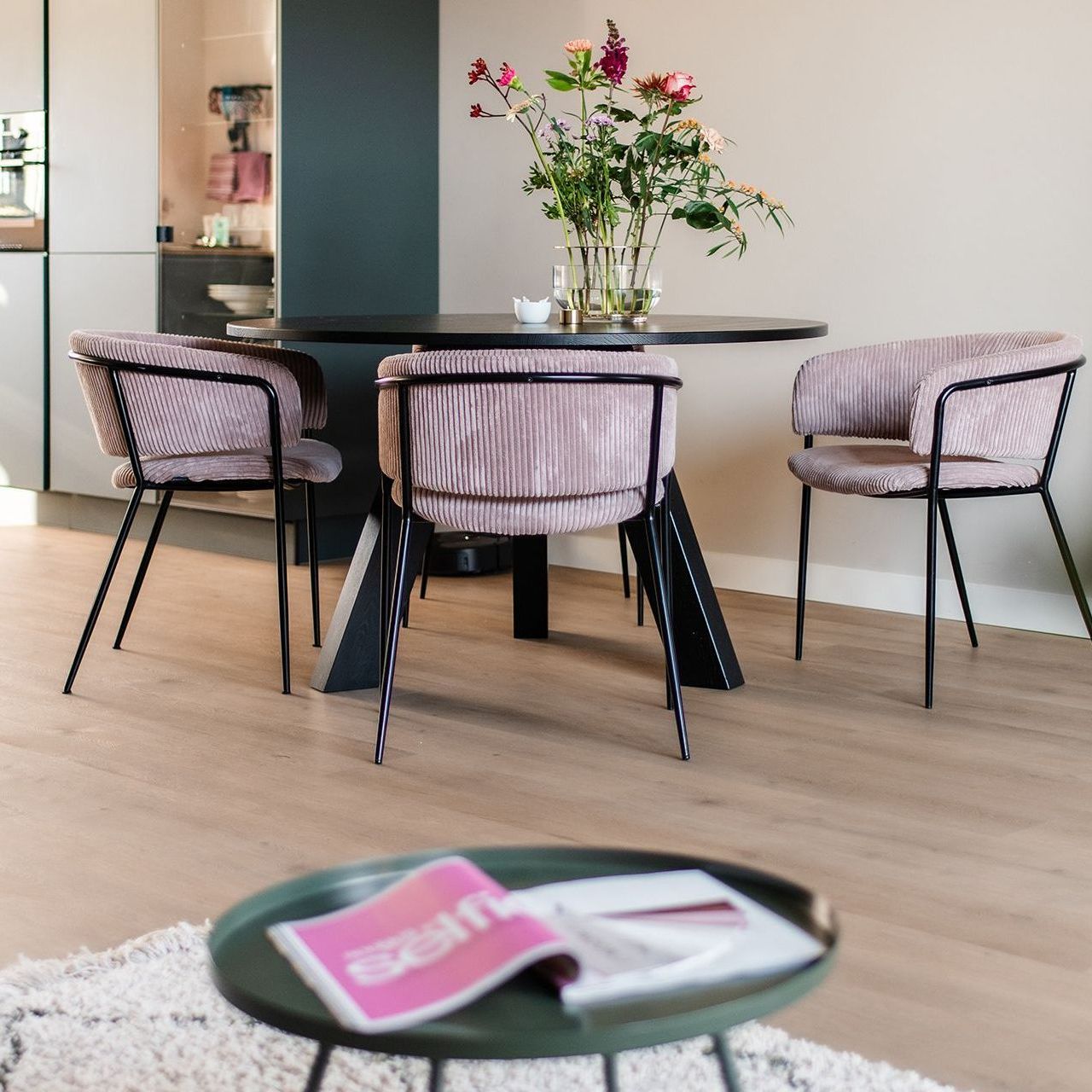 Eettafel met drie roze stoelen. Salontafel met tijdschriftenrek. Vaas met bloemen.