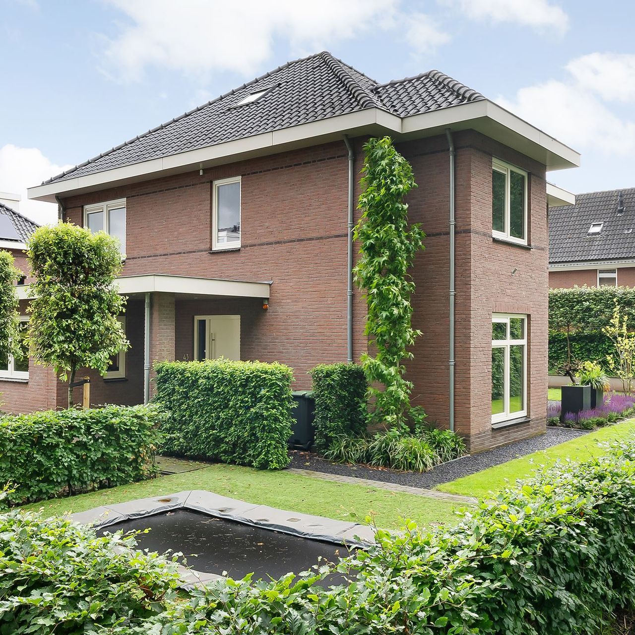 Bakstenen huis met twee verdiepingen en een voortuin. In de tuin staan ​​groene hagen en een trampoline.