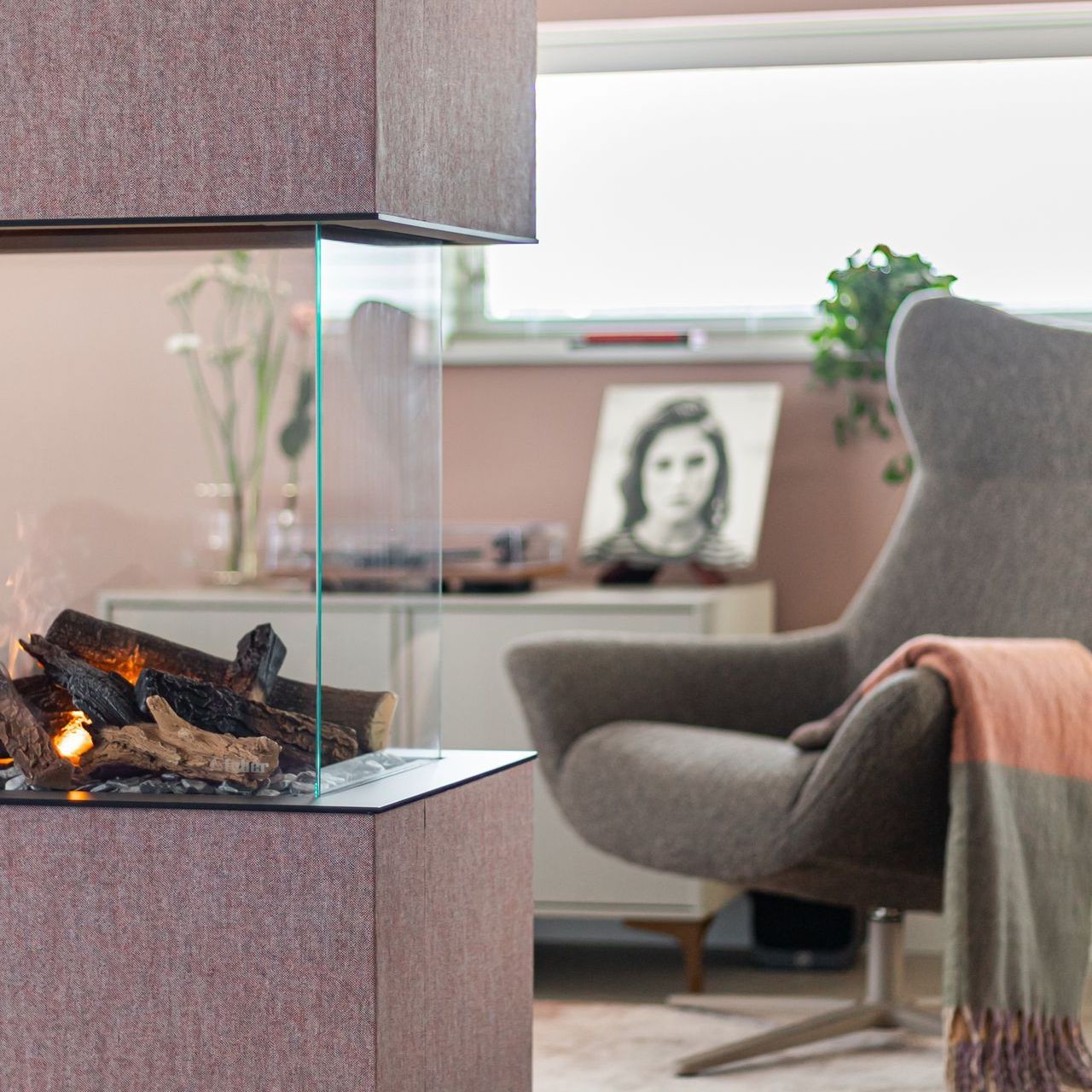 Moderne elektrische open haard met houtblokken in een woonkamer met een grijze stoel en roze muren.