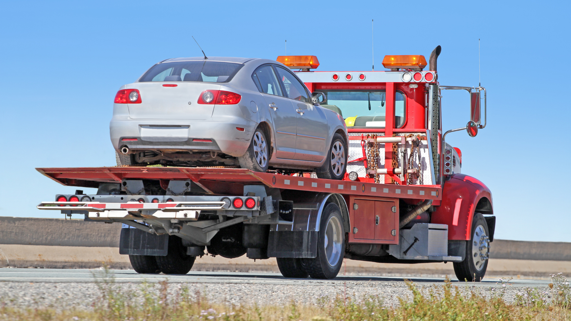 Silver car loaded on a red tow truck on a sunny road.