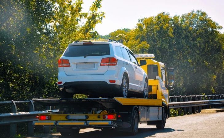 White SUV being towed by a yellow tow truck on a road with trees in the background.